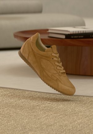 Tan suede sneaker with perforated design and flat laces. Features a rounded toe and textured rubber sole, suspended above a textured surface.