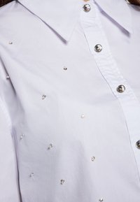 Camicia bianca con colletto classico, caratterizzata da decorazioni in tono argento sparse su tutto il tessuto e bottoni lucidi.