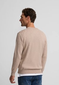 Jersey de punto beige con cuello redondo y puños acanalados, usado sobre una camisa blanca. La tela parece suave con una textura lisa.