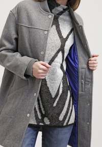 Cappotto grigio in misto lana dallo stile bomber, con bottoni a pressione argentati, polsini a coste e foderato di blu. Indossato sopra un maglione lavorato a maglia con motivo.