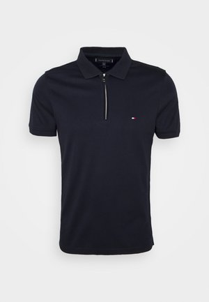Marineblaues Polohemd mit kurzen Ärmeln, Kragen, silbernem Reißverschluss vorne und kleinem roten, weißen und blauen Logo auf der Brust.