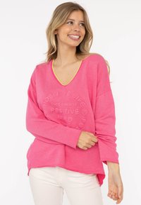 Donna sorridente che indossa un top a maniche lunghe di un brillante colore rosa, con la scritta "Vita Positiva, Buona Energia, Mente Positiva", e pantaloni bianchi, in piedi su uno sfondo bianco.