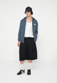 Siva zip-up dukserica, bijeli top bez rukava, crne kratke hlače, crne tenisice, bijele čarape, crna kapa. Dukserica ima grafiku i tekst na rukavima.