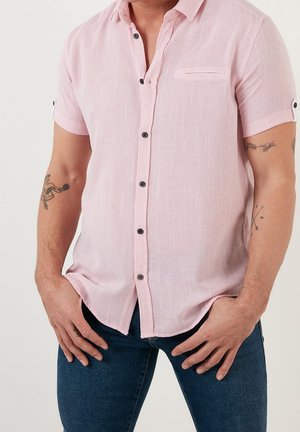 Hombre con camisa de manga corta color rosa claro abotonada y vaqueros azules, con las manos apoyadas cerca de las caderas, mostrando tatuajes en el antebrazo.