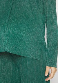 Smaragdgrön strukturerad outfit med en glittrande effekt; består av en knäppt skjorta och vida byxor som visar en flödande design och långa ärmar.