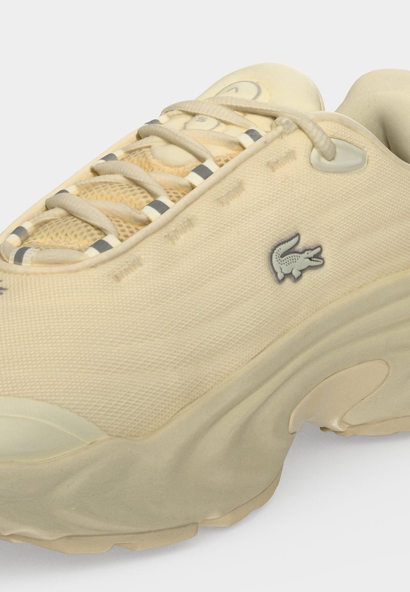 Sneaker beige con suola spessa, tomaia in mesh e tessuto, lacci beige e piccolo logo del coccodrillo sul lato.