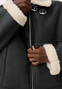 Des mains zippant une veste en cuir noir avec des poignets et un col en shearling blanc, dotée de deux boucles en argent au niveau du cou.