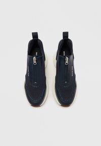 Μπλε ναυτικού slip-on αθλητικά παπούτσια με ύφασμα από δίχτυ, δερμάτινες λεπτομέρειες και ασημένιο φερμουάρ. Η σόλα είναι λευκή με ανοιχτά καφέ κάτω μέρος.