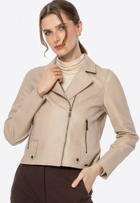 Beige leren motorjack met een gekanteld kraag, een voorrits en twee ritssluitingen. Korte stijl met een soepele textuur.