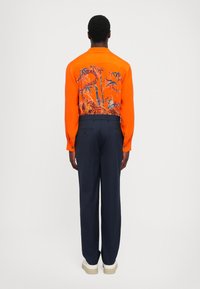 Oranssi pitkähihaisen paidan, jossa on sininen kukkakuosi takana, yhdistettynä tummansinisiin housuihin ja valkoisella pohjalla varustettuihin kenkiin.