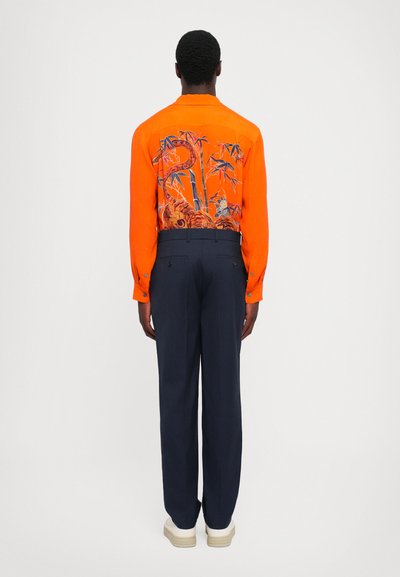 Orange langærmet skjorte med et blåt blomstermønster på ryggen, parret med marineblå bukser og sko med hvid sål.