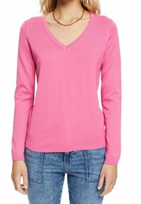 Różowy sweter z dekoltem w serek i długimi rękawami, wykonany z miękkiej dzianiny. Charakteryzuje się prostym designem oraz ściągaczami przy mankietach i na dole. Noszony na niebieskich dżinsach.