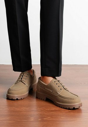 Chaussures à lacets en daim marron pour homme avec semelles épaisses en caoutchouc portées avec un pantalon noir sur un sol en bois.