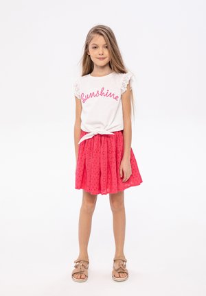Valkoinen T-paita, jossa on vaaleanpunaista "Sunshine"-tekstiä, pitsireunat hihansuissa, yhdistettynä vaaleanpunaiseen skater-hameeseen, jossa on silppukuvio ja beigeet sandaalit.
