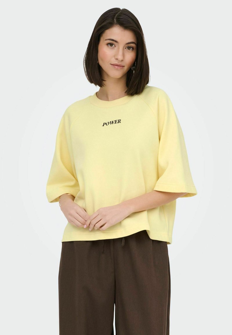 Femme aux cheveux foncés mi-longs portant une chemise ample jaune pâle avec le texte "POWER" et un pantalon large sombre, debout devant un fond uni.