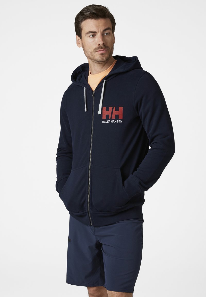 Sudadera con cremallera en color navy, fabricada en un material suave, con cordones grises. Presenta un logo en rojo y blanco de "Helly Hansen" en el pecho.