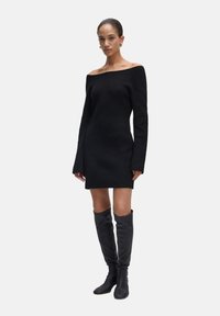 Schwarzes schulterfreies Kleid mit gerippter Textur, langen Ärmeln und taillierter Passform. Kombiniert mit kniehohen schwarzen Stiefeln.