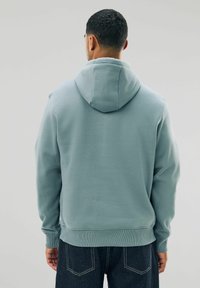 Uomo che indossa una felpa azzurro chiaro e jeans scuri, in piedi con la schiena rivolta verso la fotocamera su uno sfondo neutro.