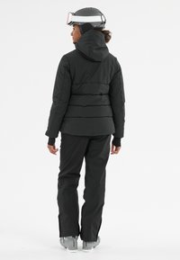 Zwarte pufferjack met capuchon, met een gestructureerd ontwerp en een aangesloten vorm, gecombineerd met zwarte sneeuwbroeken en grijze skibotten. Inclusief goggles.