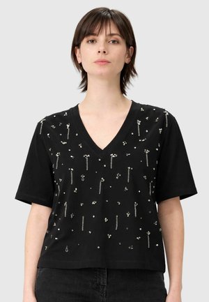 Donna che indossa una maglietta nera a scollo a V con maniche corte decorata con piccole perline d'argento e ornamenti simili a catene, abbinata a pantaloni scuri.