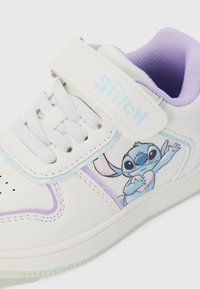 Baskets blanches pour enfants avec des bordures et un revêtement violet, mettant en avant Disney Stitch tenant un cœur sur le côté et un logo "Stitch" sur la sangle.