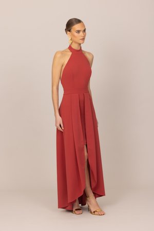 Žena v červenom šatách bez rukávov s vysokým golierom a predným rázporkom, obutá v zlatých sandáloch na remienkoch, stojaca pred jednoduchým béžovým pozadím.