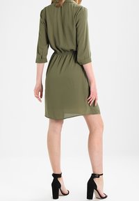 Oliwkowa sukienka koszulowa z rękawami trzy czwarte, talią podkreśloną paskiem oraz gładkim materiałem; zestawiona z czarnymi szpilkami na kostkę.