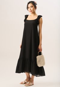Black dress with ruffled shoulder straps, a square neckline, and a flared hem. Model holds a woven beige bag and wears light sandals.