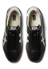 Černé kožené tenisky s bílými pruhy a tkaničkami, se vyztuženým interiérem a hnědou gumovou podrážkou. Viditelné branding Onitsuka Tiger.