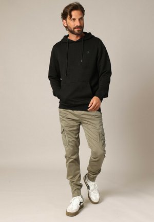 Haut de hoodie noir texturé avec cordons de serrage, pantalon cargo olive avec poches latérales et baskets blanches avec accents marron.