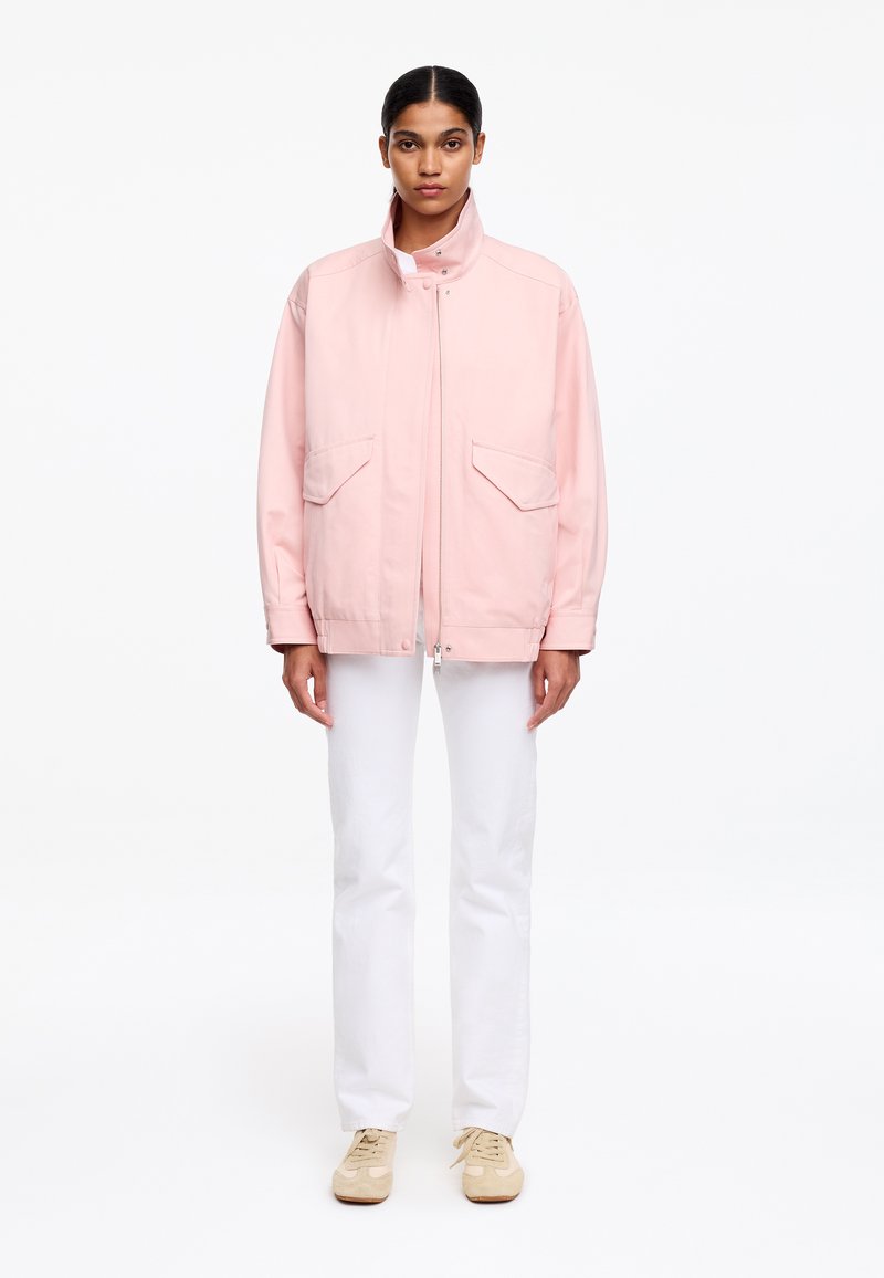 Femme debout portant une veste zippée rose clair avec poches à rabat, un pantalon blanc et des baskets beiges sur un fond blanc uni.