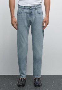 Light blue denim jeans with a straight fit, featuring front pockets and a button closure, paired with black loafers on a gray background.