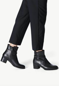 Bottines en cuir noir avec une fermeture éclair sur le côté, un tige effilée et un talon bloc, comportant un accent de sangle décoratif près du haut.