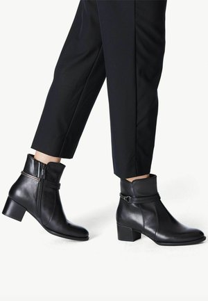 Botas de tobillo de cuero negro con cremallera lateral, caña cónica y tacón cuadrado, con un detalle decorativo en forma de correa cerca de la parte superior.