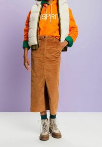 Brauner Cord-Midi-Rock mit vorderem Schlitz, kombiniert mit einem orangefarbenen Hoodie und einer grünen Weste. Abgerundet mit beigen Stiefeln und grünen Socken.