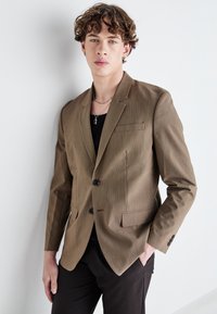 Brauner strukturierter Blazer mit Reverskragen, Ein-Knopf-Verschluss und zwei vorderen Taschen, kombiniert mit einem schwarzen Oberteil und dunklen Hosen.