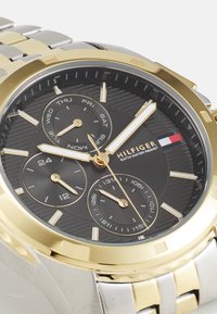 Montre chronographe avec un cadran noir texturé, un boîtier en acier inoxydable doré et argenté, trois sous-cadrans et un affichage de la date.