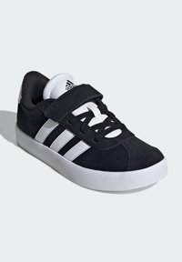 Zwarte suède sneaker met een witte rubberen zool, zwarte veters en een klittenbandsluiting. Drie witte strepen aan de zijkant.