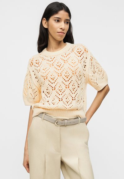 Femme portant un pull en maille crème à manches courtes avec des motifs en losange et un pantalon beige taille haute avec une ceinture claire, posant avec une main sur la hanche.