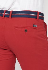 Pantalon en coton rouge à coupe ajustée, orné d'une ceinture tissée bleu marine avec des rayures blanches et rouges, et d'une poche arrière avec fermeture à bouton.