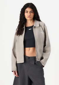 Light gray zip-up jacket with a collar, featuring embroidered "Obey" logo, worn over a black crop top. Textured fabric with relaxed fit.