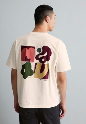 Mann, der mit dem Rücken zum Betrachter steht, trägt ein beigefarbenes T-Shirt mit bunten abstrakten Formen in vier Quadraten auf dem Rücken, kombiniert mit schwarzen Hosen.