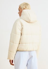 Casaco acolchoado de cor creme com capuz, apresentando um acolchoamento horizontal e punhos canelados. Superfície texturizada com um acabamento suave.