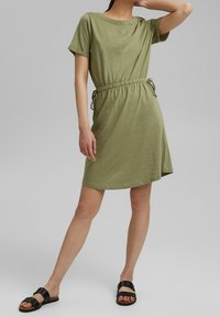 Robe t-shirt vert olive à coupe décontractée et manches courtes, resserrée à la taille avec des cordons réglables. Associée à des sandales noires.