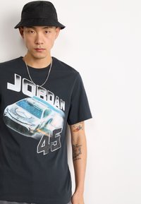 Camiseta negra con un gráfico de un coche de carreras y el texto "JORDAN 45" en letras blancas en negrita. El modelo lleva un sombrero tipo cubo negro.