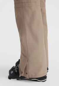 Light brown snow pants with a straight cut, featuring side vents and a smooth texture. Paired with black ski boots.