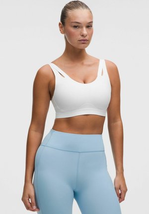Vrouw met haar naar achteren gebonden, gekleed in een witte sportbeha met dubbele bandjes en lichtblauwe leggings met hoge taille tegen een effen achtergrond.