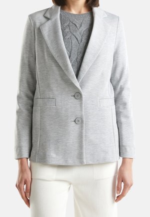 Femme portant un blazer gris clair à deux boutons sur un pull en maille torsadée gris et un pantalon blanc sur un fond uni.