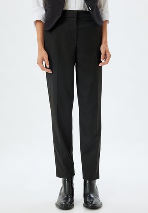 Pantalones negros a medida con corte recto, textura suave y cinturilla de altura media. Llevados con botines negros brillantes.