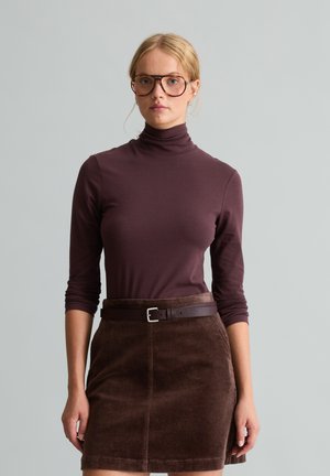 Kobieta o blond włosach, nosząca duże okulary, ciemnobrązowy golf i brązową spódnicę sztruksową z paskiem, stojąca na tle jasnoszarym.
